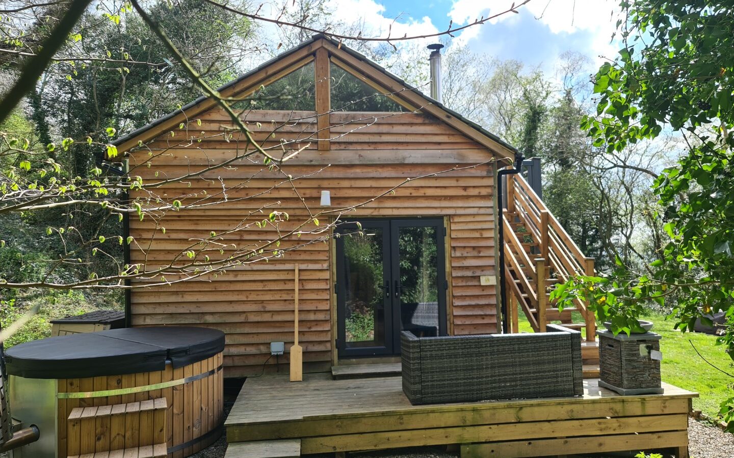 A charming wooden cabin with a treehouse vibe features a deck, outdoor seating, and a hot tub enveloped by lush greenery. Perfect for stargazing, stairs lead to an inviting upper level that promises tranquility and comfort.