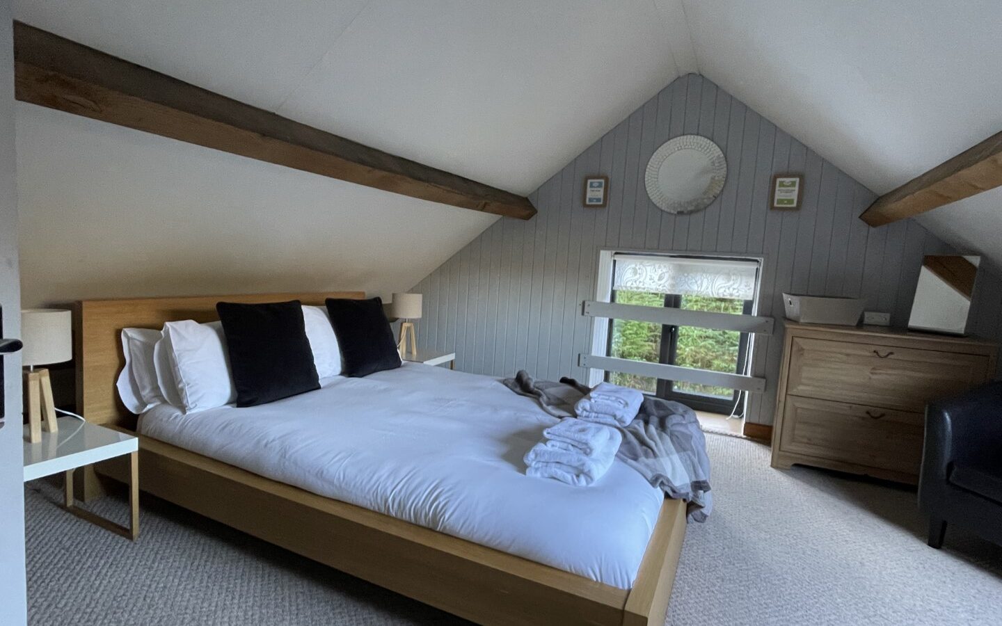 A cozy attic bedroom in Trallwm Forest Cottages features a wooden bed, white bedding, and black pillows. A dresser and mirror rest against a wood-paneled wall, creating a rustic charm.