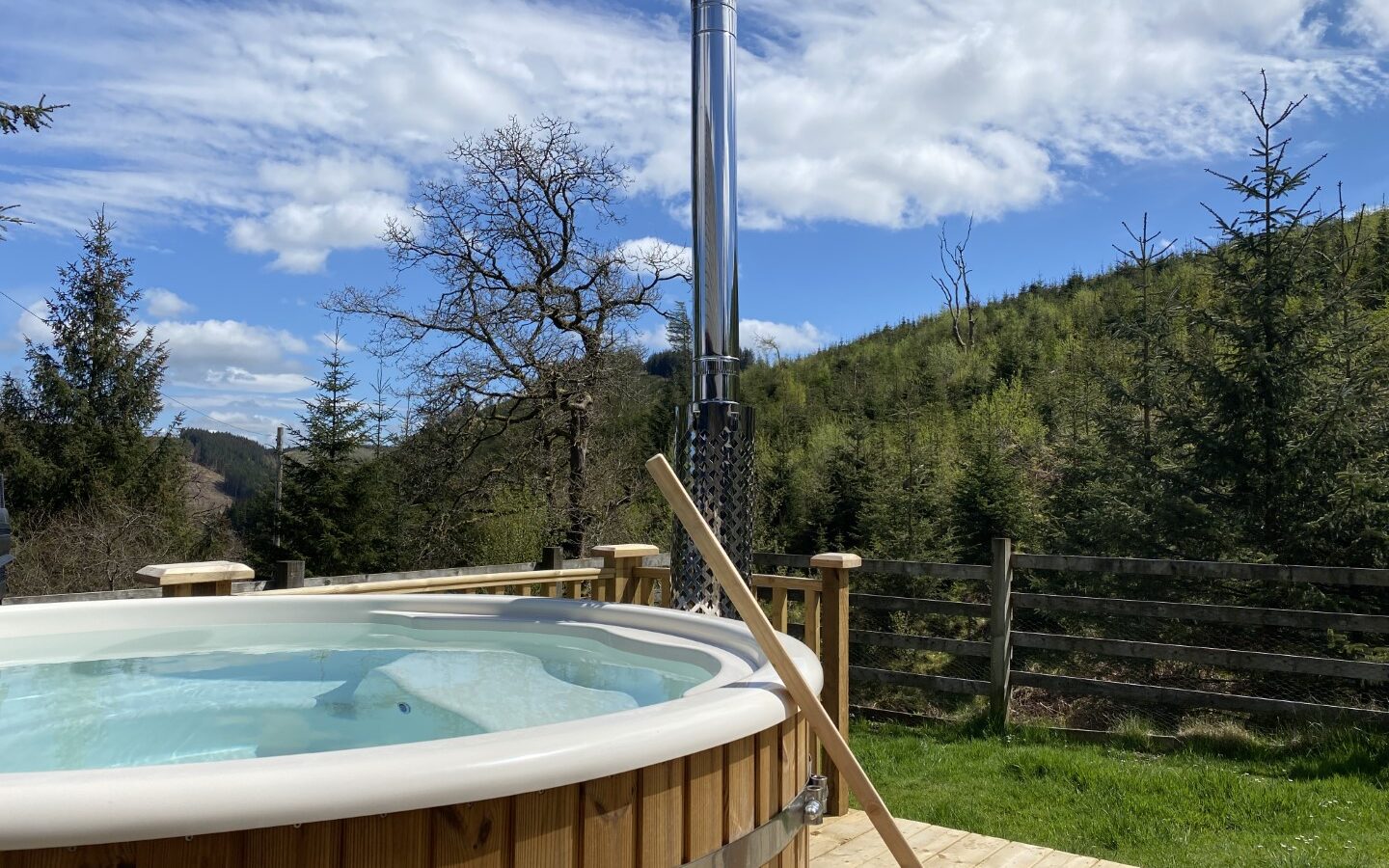 A wooden hot tub with a chimney stands on a deck at Trallwm Forest Cottages, surrounded by trees under a partly cloudy sky.