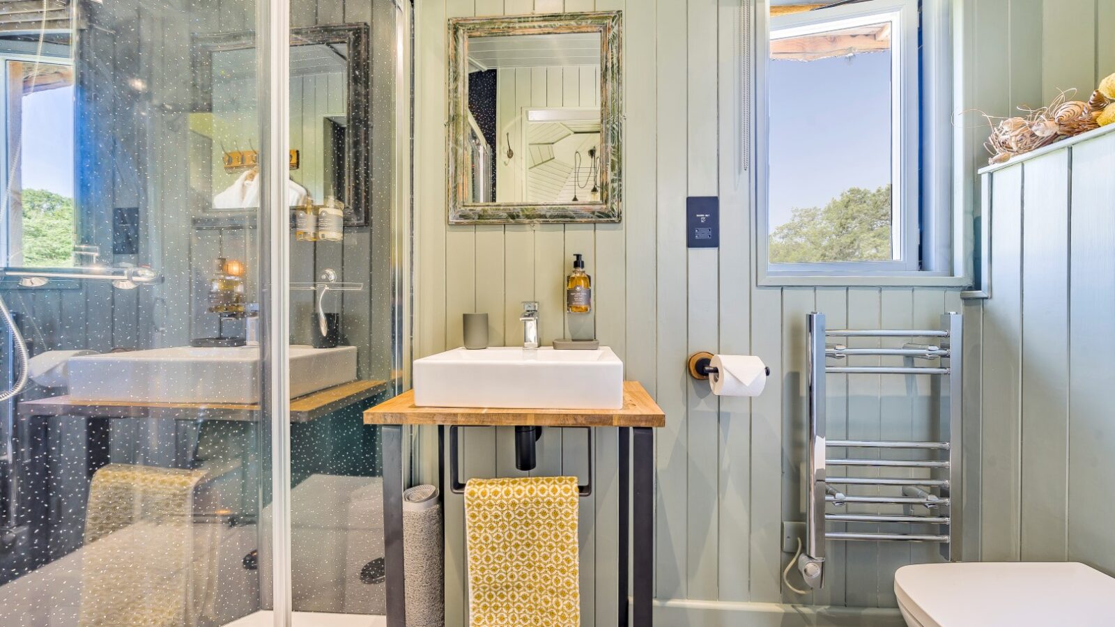 A modern bathroom with light green paneled walls. It features a glass-enclosed shower, a white sink with a wooden countertop, and a wall-mounted mirror—all elements reminiscent of Erwain Escapes' cozy cabins. A towel rack with a yellow towel and a window with striped blinds complete the serene space.