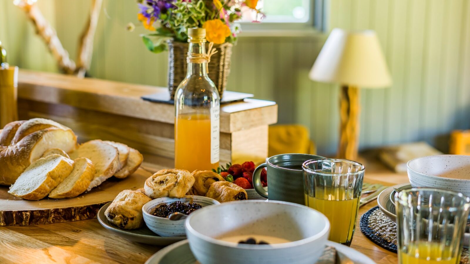 A wooden table set with breakfast items including slices of bread, pastries, bowls filled with yogurt or cereal, glasses of orange juice, a bottle of orange juice, and a bouquet of flowers in a vase. A window and a lamp are visible in the cozy cabin backdrop from Erwain Escapes.