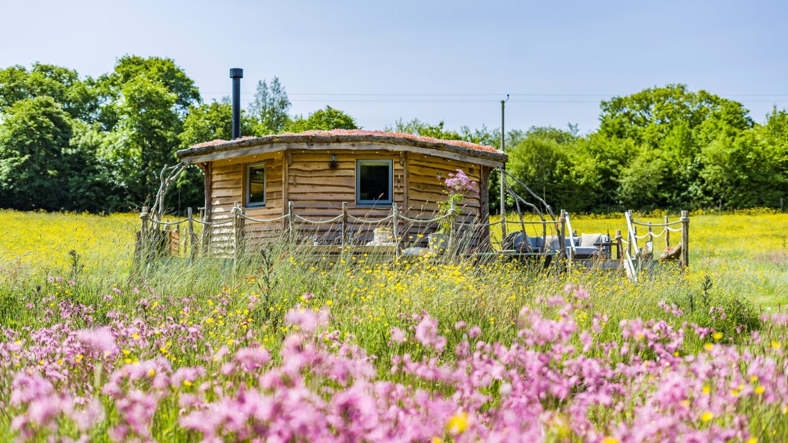 Erwain Escapes offers The Cabin, a small, round wooden sanctuary nestled in a vibrant meadow filled with pink and yellow wildflowers. Enclosed by a charming rope fence, it's set against a backdrop of lush green trees under a bright blue sky.