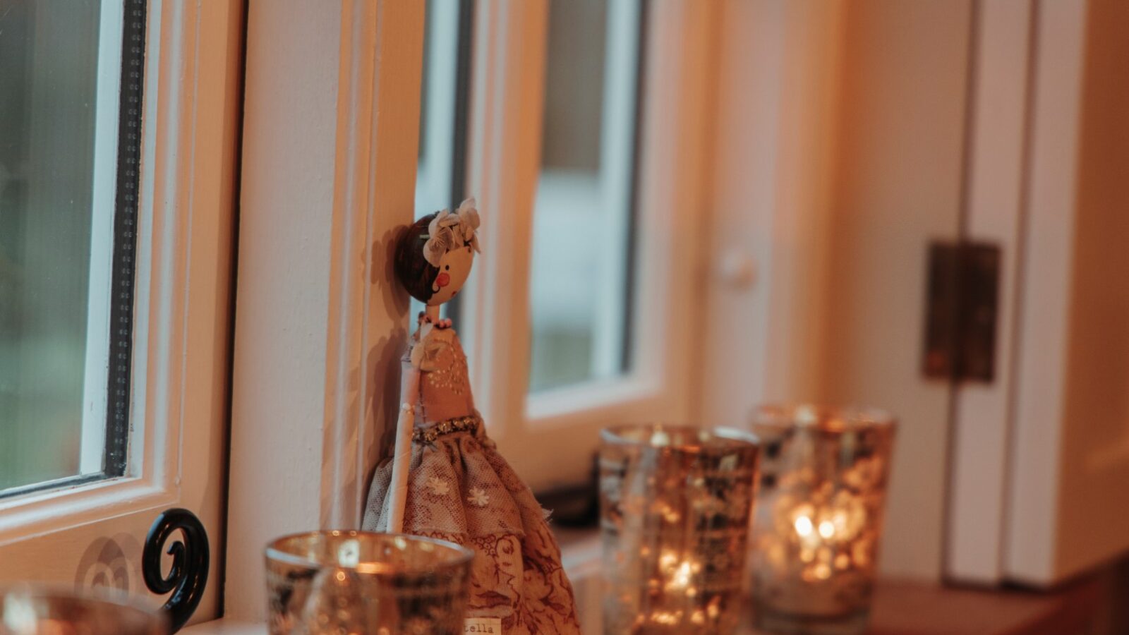 Decorative candles and a small fabric doll are placed on a windowsill, casting a warm glow in a softly lit room reminiscent of a cozy garden hut.