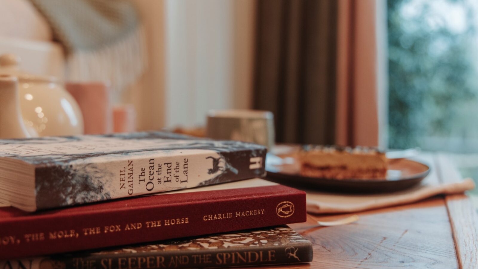 A stack of books on a wooden table alongside a teapot and a plate with a slice of cake, with the blurred window revealing hints of the garden beyond.