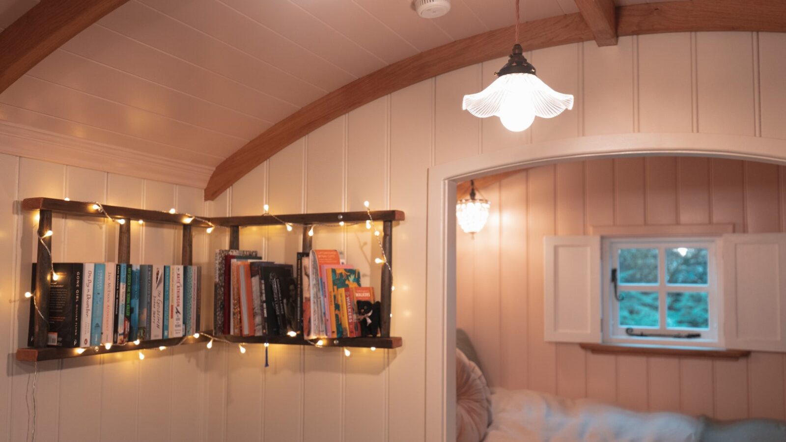 Cozy room resembling a charming hut, with a bookshelf, string lights, and a bed. Soft lighting and a small window by the bed overlook the tranquil garden, creating a warm atmosphere.