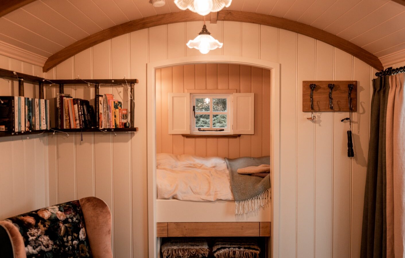 The cozy interior of this tiny house feels like a welcoming garden retreat, featuring a snug bed nook, bookshelves, an inviting armchair, and handy wall hooks all bathed in warm lighting.