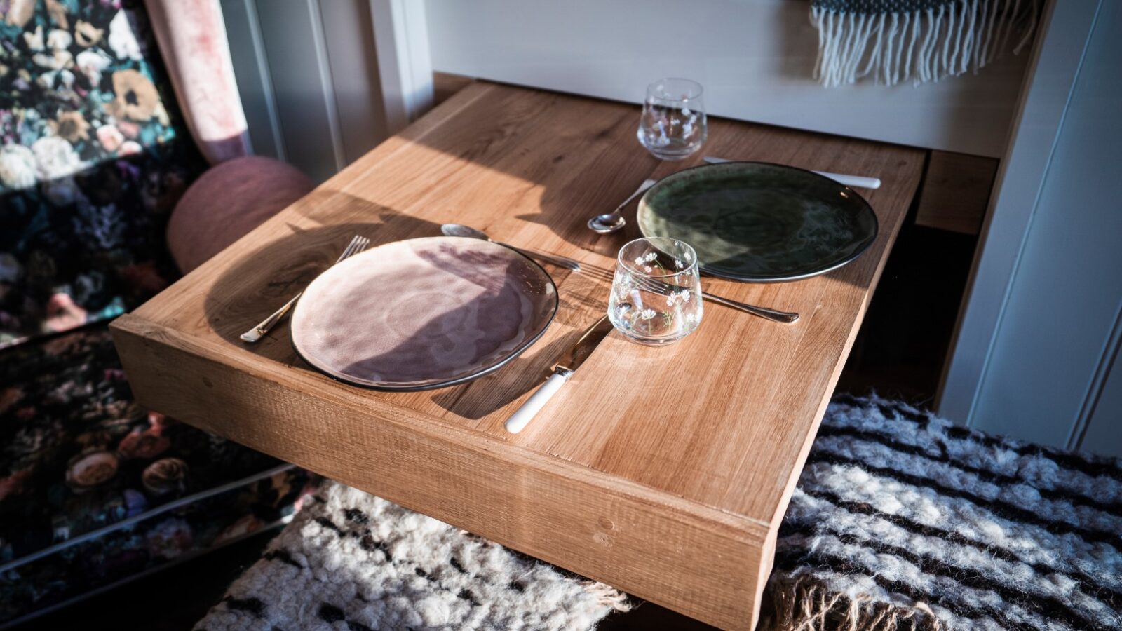 A wooden table set for two with glassware, cutlery, and green and pink plates sits charmingly in the Garden Hut, surrounded by a bench and cushioned seating. This cozy setup blends seamlessly with the enchanting outdoor decor.