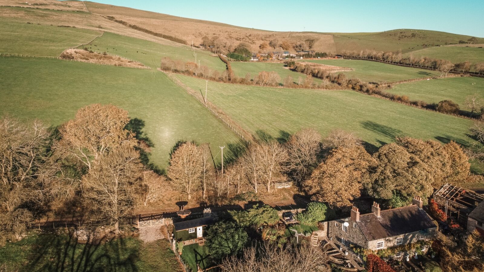 An aerial view of a rural landscape reveals fields, trees, and a charming house with a lush garden. A quaint hut nestles among the greenery, while hills stand majestically in the distance under a clear blue sky.