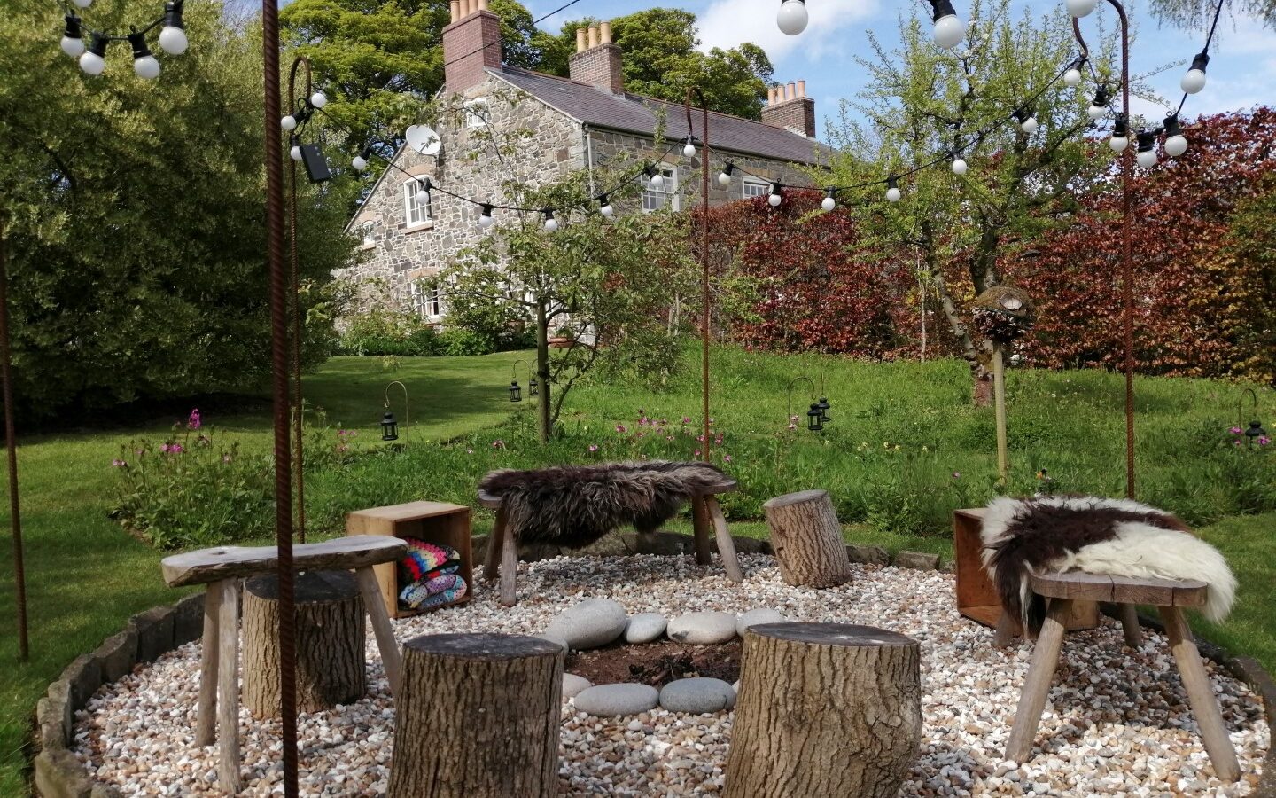 The garden features an inviting outdoor seating area with log stools and plush fur throws around a fire pit. Surrounded by twinkling string lights, the scene is idyllic, complemented by a cozy hut nestled in the background.