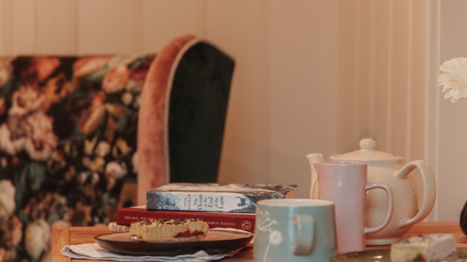 A cozy setting inside a quaint hut features books, a teapot, a mug, and plates of cake on a wooden table, next to a floral-patterned chair that offers views of the garden.