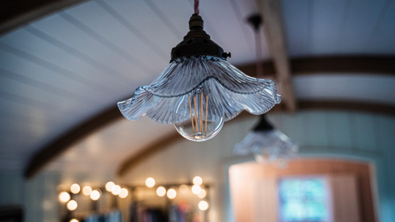 Close-up of a decorative pendant light with a fluted glass shade, set against a blurred background reminiscent of a cozy garden hut, with wooden beams and string lights adding charm.