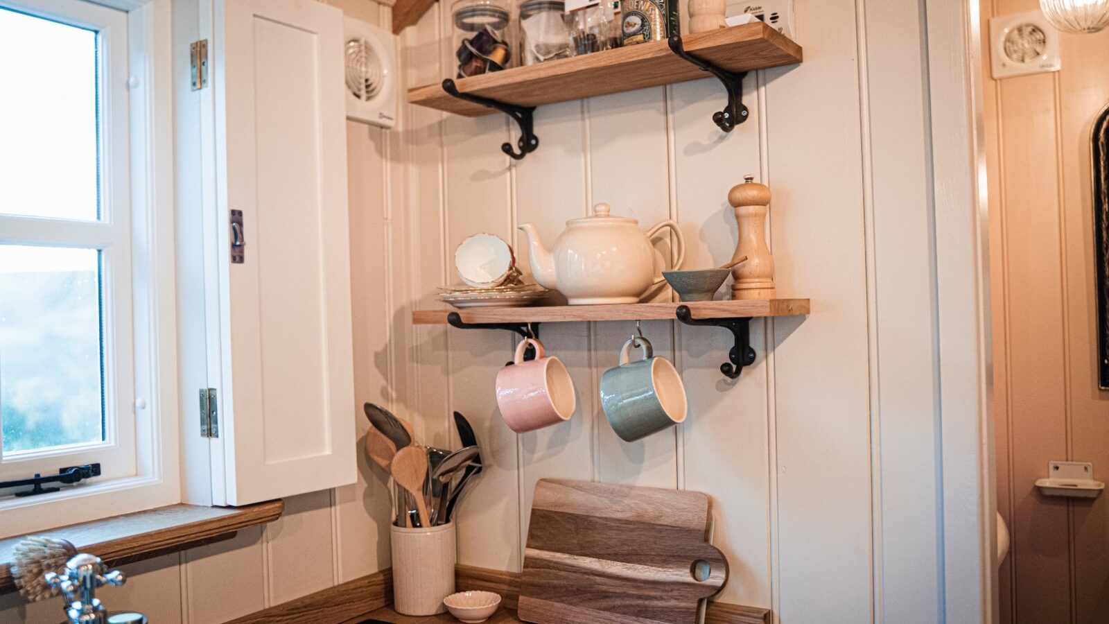 Cozy kitchen corner reminiscent of a quaint hut, with wooden shelves holding jars, a teapot, mugs, utensils, a cutting board, and a pepper grinder. This delightful nook brings the charm of the garden indoors.