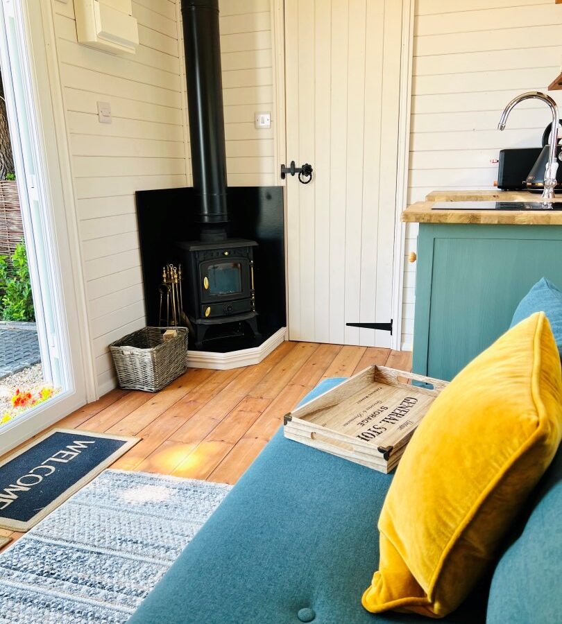 A cozy interior features a teal couch with mustard yellow pillows, a wooden tray on the seat, and a rustic wood-burning stove in the corner. In The Green Hut, a door with a black handle is in the background next to a small kitchen area with a counter. A welcome mat is near the entrance.