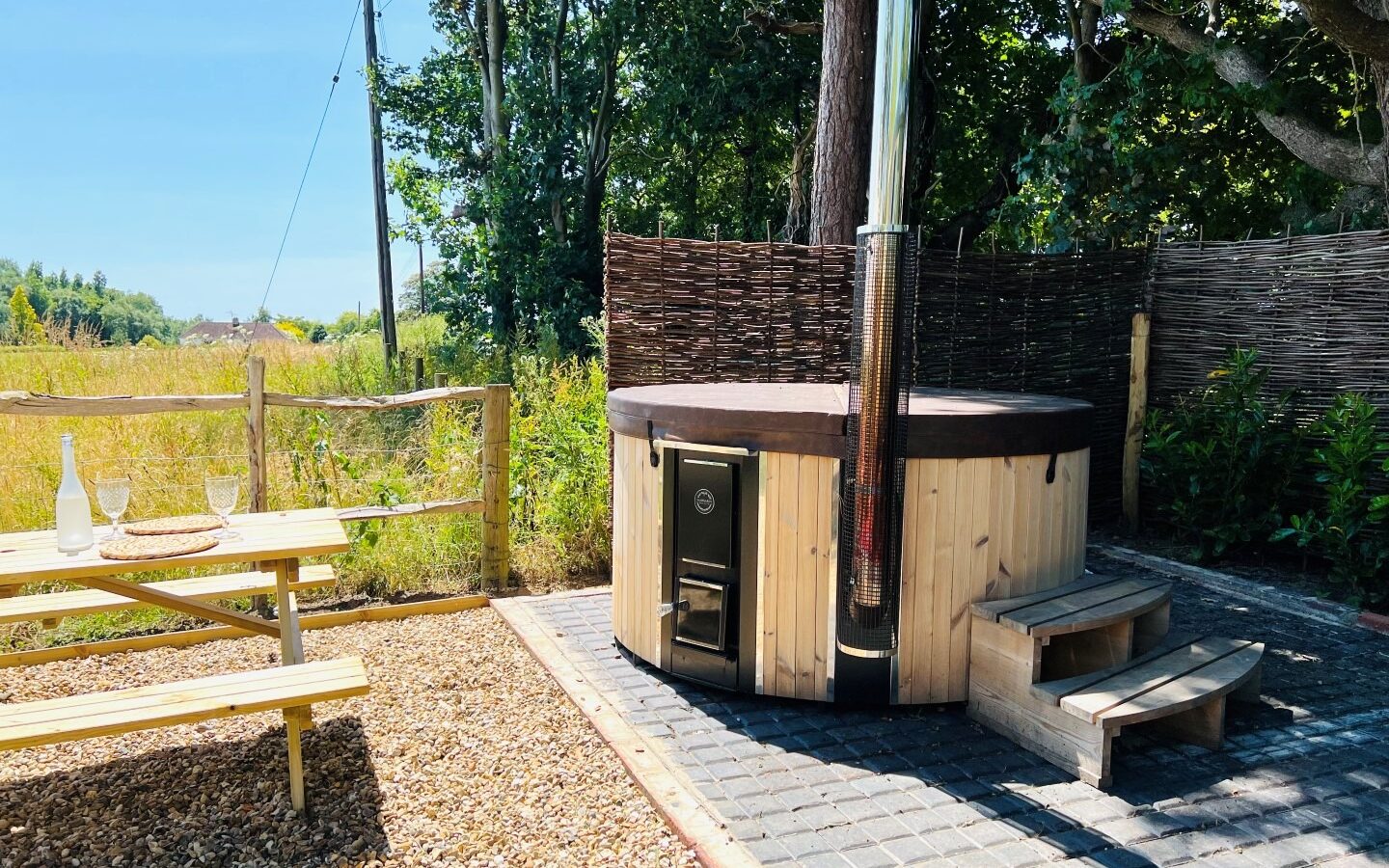 A sunlit outdoor area with a wooden hot tub and steps, surrounded by a wicker privacy screen and trees. A picnic table sits nearby on a gravel area with a bottle and two glasses on the table. The background has a grassy field, clear blue sky, and The Green Hut.