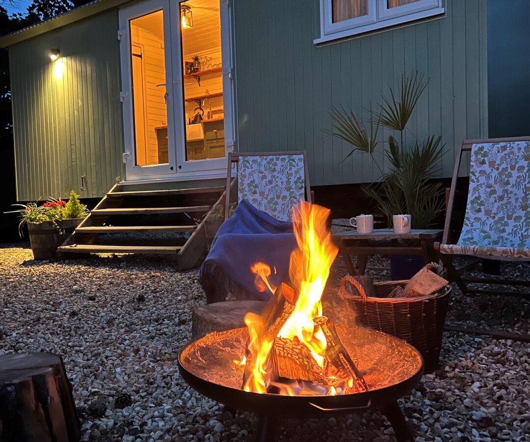 A cozy nighttime scene with a campfire burning in a fire pit in the foreground. Two chairs are positioned nearby, one draped with a blanket. The Green Hut, a small illuminated cabin with an inviting interior, is in the background, surrounded by trees and outdoor lights.