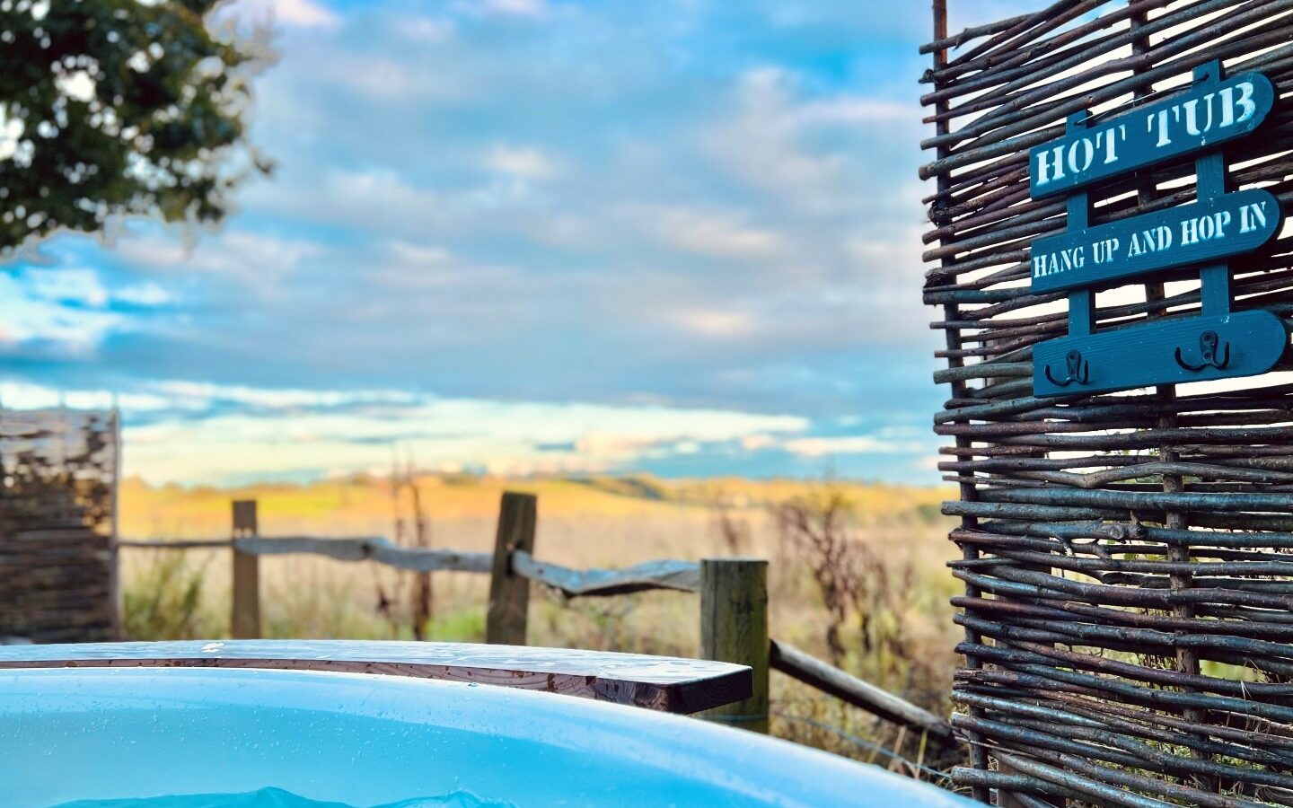 A hot tub with blue water sits outdoors under a clear sky with scattered clouds. Nearby, a Green Hut stands behind a wooden fence and a sign reading 