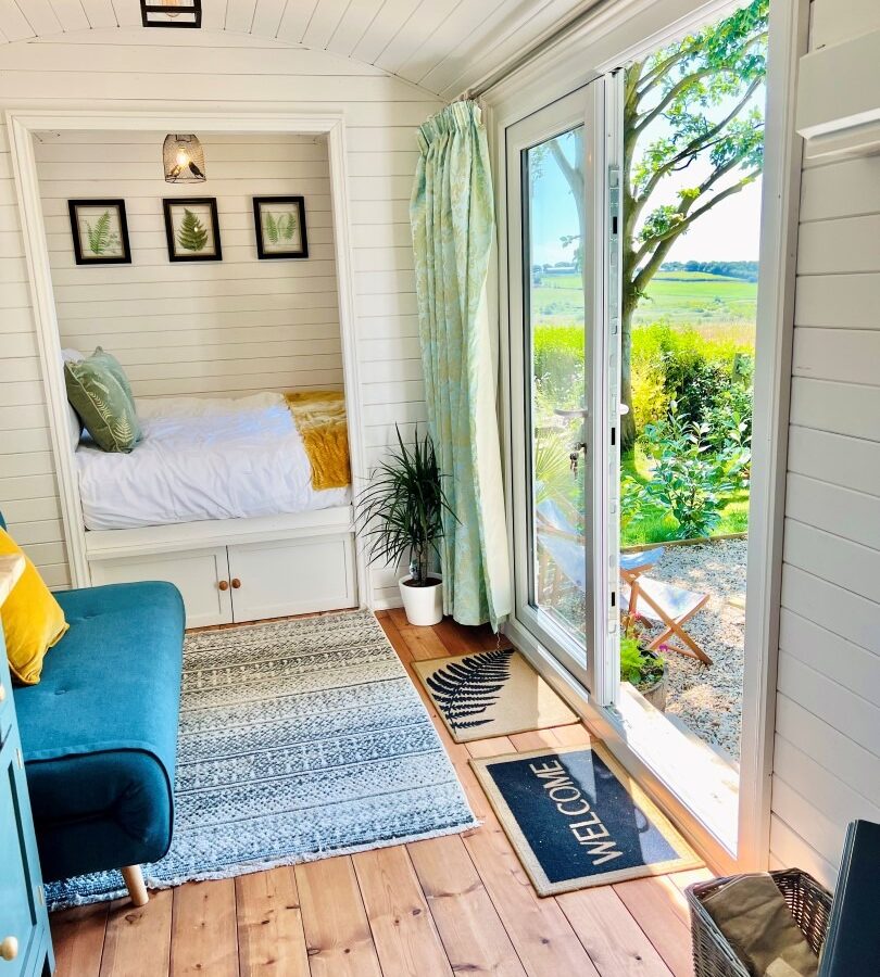 The Green Hut offers a cozy, brightly lit tiny home interior with white paneled walls and wood flooring. Inside, there is a bed nook with framed plant art above, a blue couch with yellow pillows, and an open glass sliding door showing a sunny outdoor view with green fields. A welcome mat sits by the door.