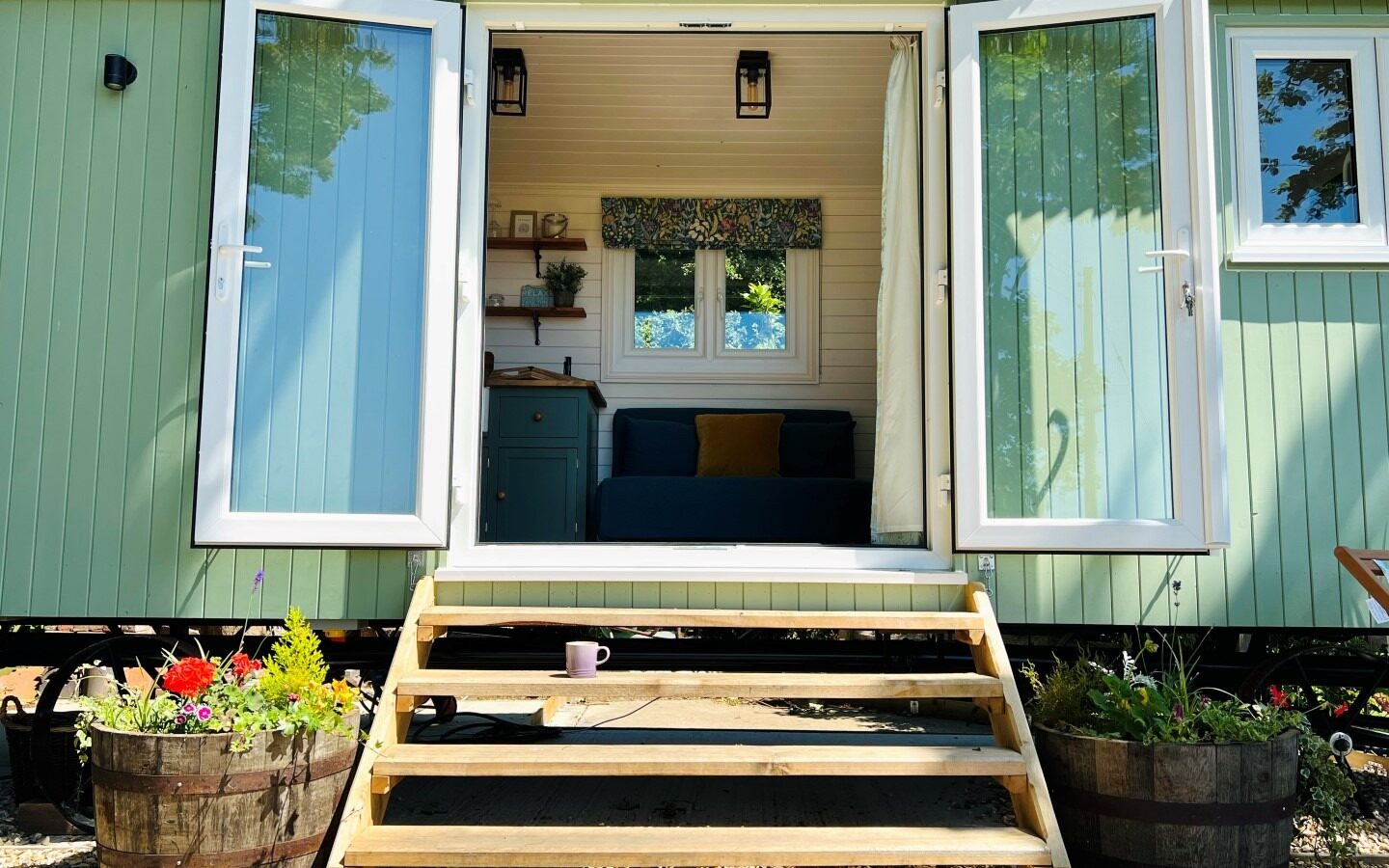 A mint green tiny house, aptly named Green Hut, with white trim features open double doors revealing a cozy interior. A wood-framed porch leads up to the entrance, flanked by two potted flower barrels. Inside, a couch and kitchen cabinets are visible. A pink mug sits on the steps of this eco-friendly retreat.