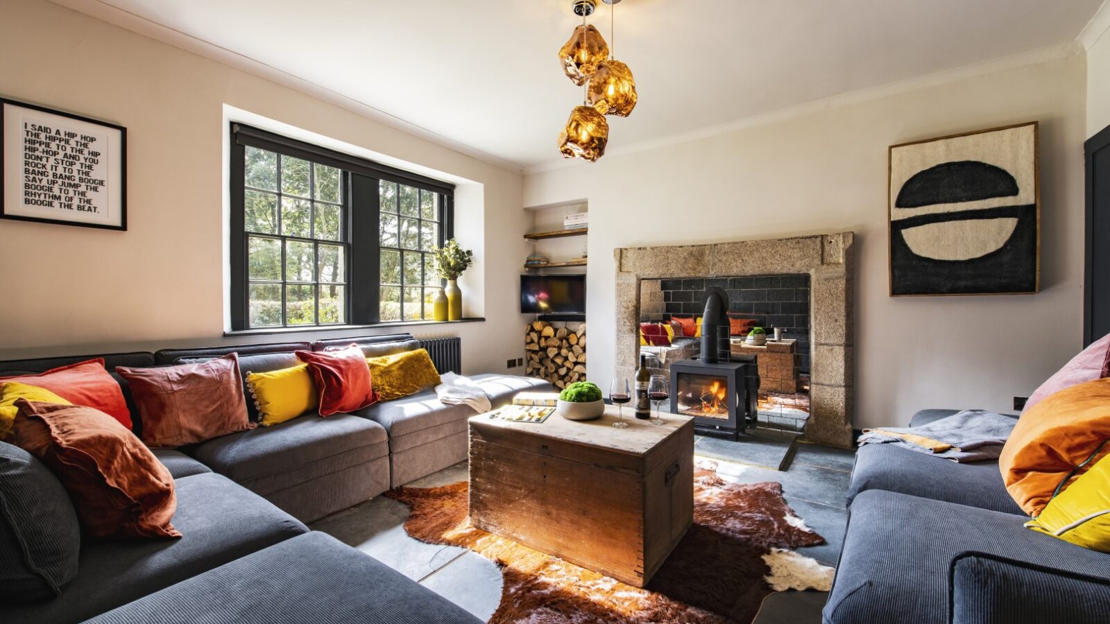 A cozy Bogee Farm living room features a sectional sofa adorned with colorful cushions. A wooden coffee table and cowhide rug sit in the center, complemented by a wood-burning stove set in the stone fireplace. Large windows embrace natural light, while a modern pendant light hangs above.