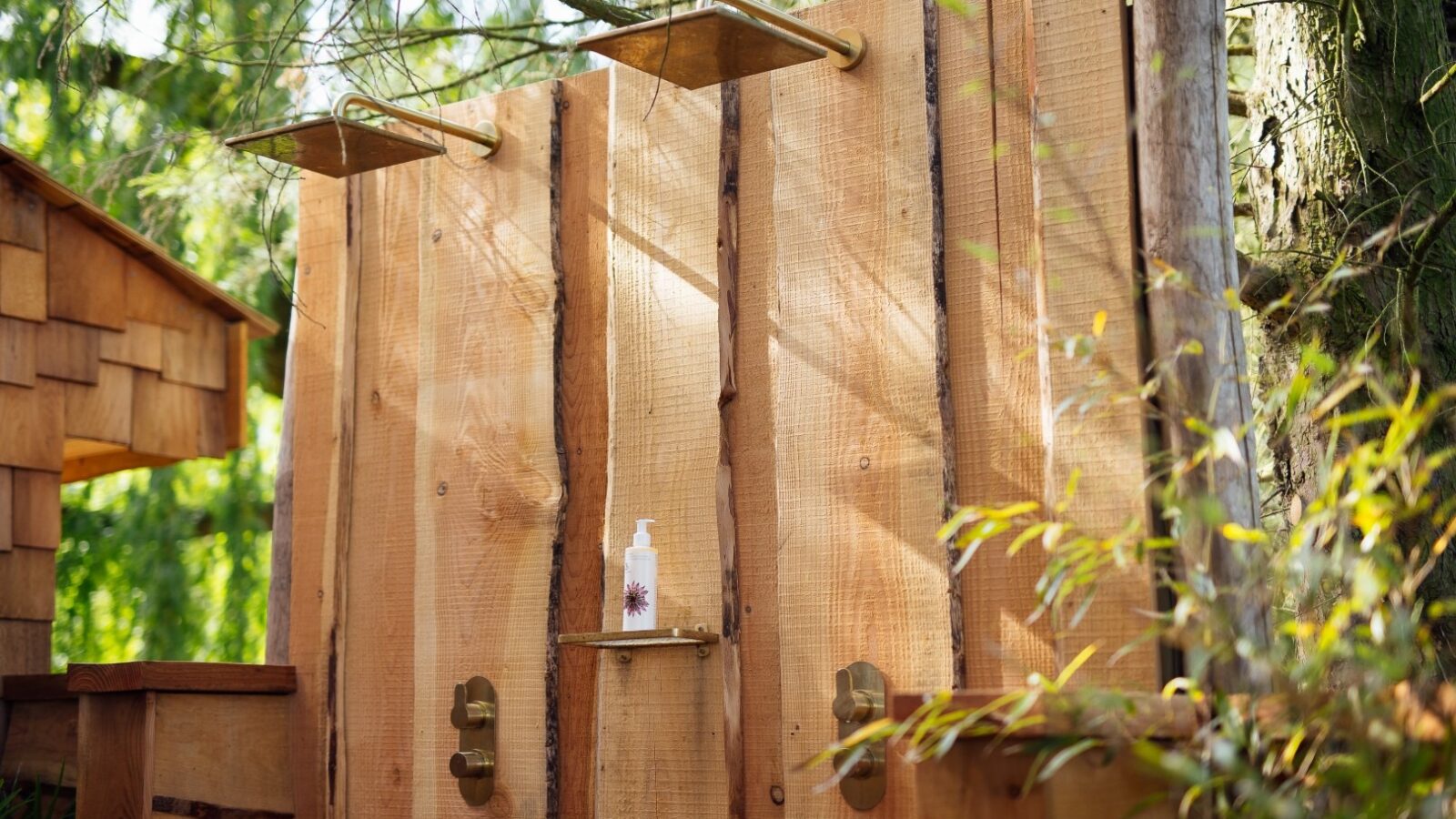 An outdoor wooden shower with two showerheads and a rustic design, surrounded by lush greenery. A small shelf between the showerheads holds a bottle of liquid soap. The shower at Fenny Castle is set under tree branches, partially shaded by a nearby structure, adding to The Orchard's charm.