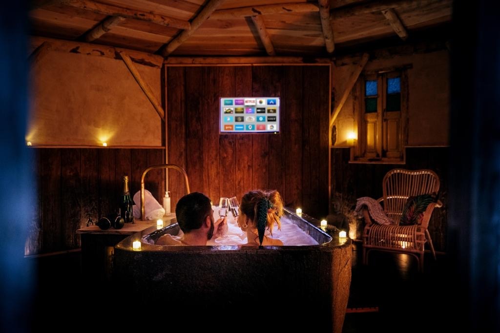 A couple relaxes in a candle-lit wooden bathtub inside a cozy, rustic room at The Orchard, with wooden walls and a beamed ceiling. They hold wine glasses as they enjoy the ambiance, while a television screen displaying various apps is mounted on the wall. A wicker chair and a window are seen in the background.