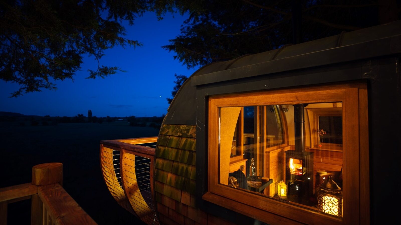 A cozy cabin with warm interior lights is nestled among trees at dusk near Fenny Castle. The cabin features a large window revealing a softly lit room with wooden furnishings and a lantern, set against a dark blue sky. The balcony, visible with wooden railings, overlooks the serene beauty of The Orchard.