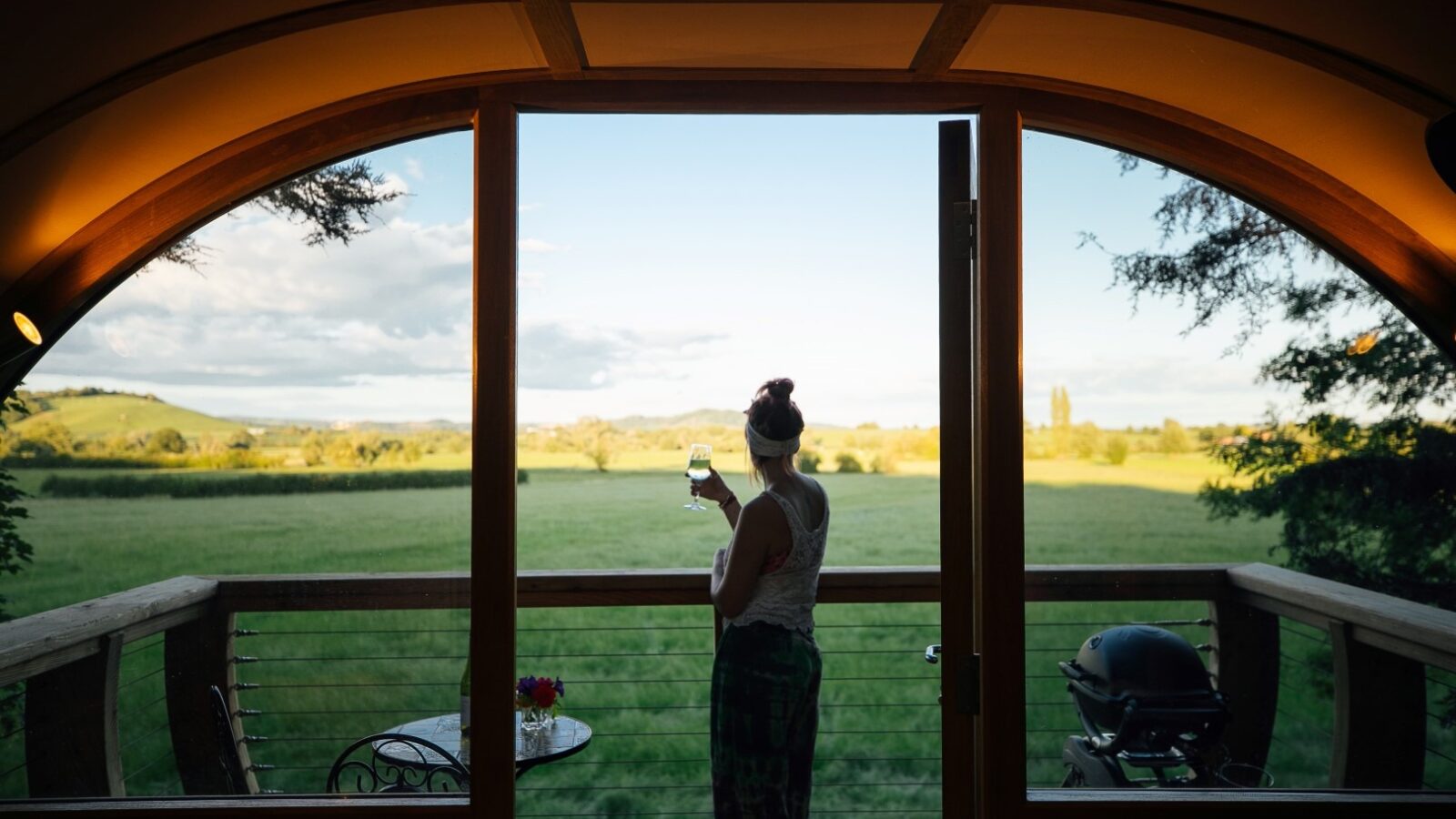 A person stands on a wooden deck holding a glass, overlooking a vast scenic landscape with green fields and distant hills on a clear day. The deck at The Orchard features flowers, chairs, and a barbecue grill. The serene view through the arched doorway adds charm to the setting near Fenny Castle.