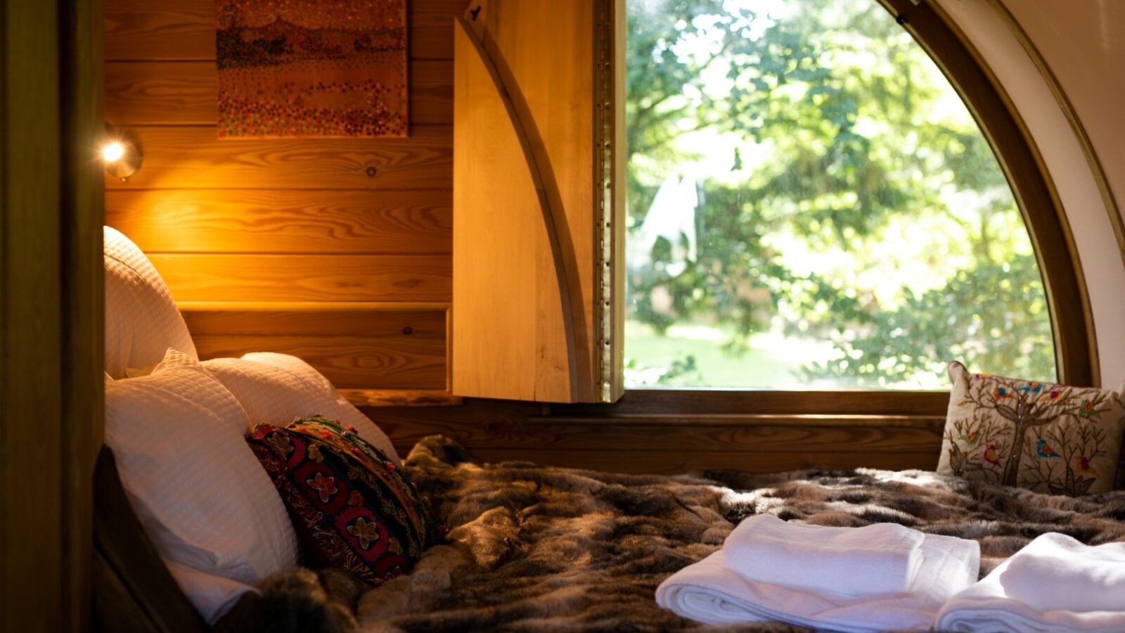 A cozy wooden cabin bedroom with a large circular window overlooking The Orchard, a green, sunlit garden. The bed is covered with a fur blanket, white pillows, and colorful throw pillows. A small towel set is neatly folded on the bed, and a painting of Fenny Castle decorates the wooden wall.