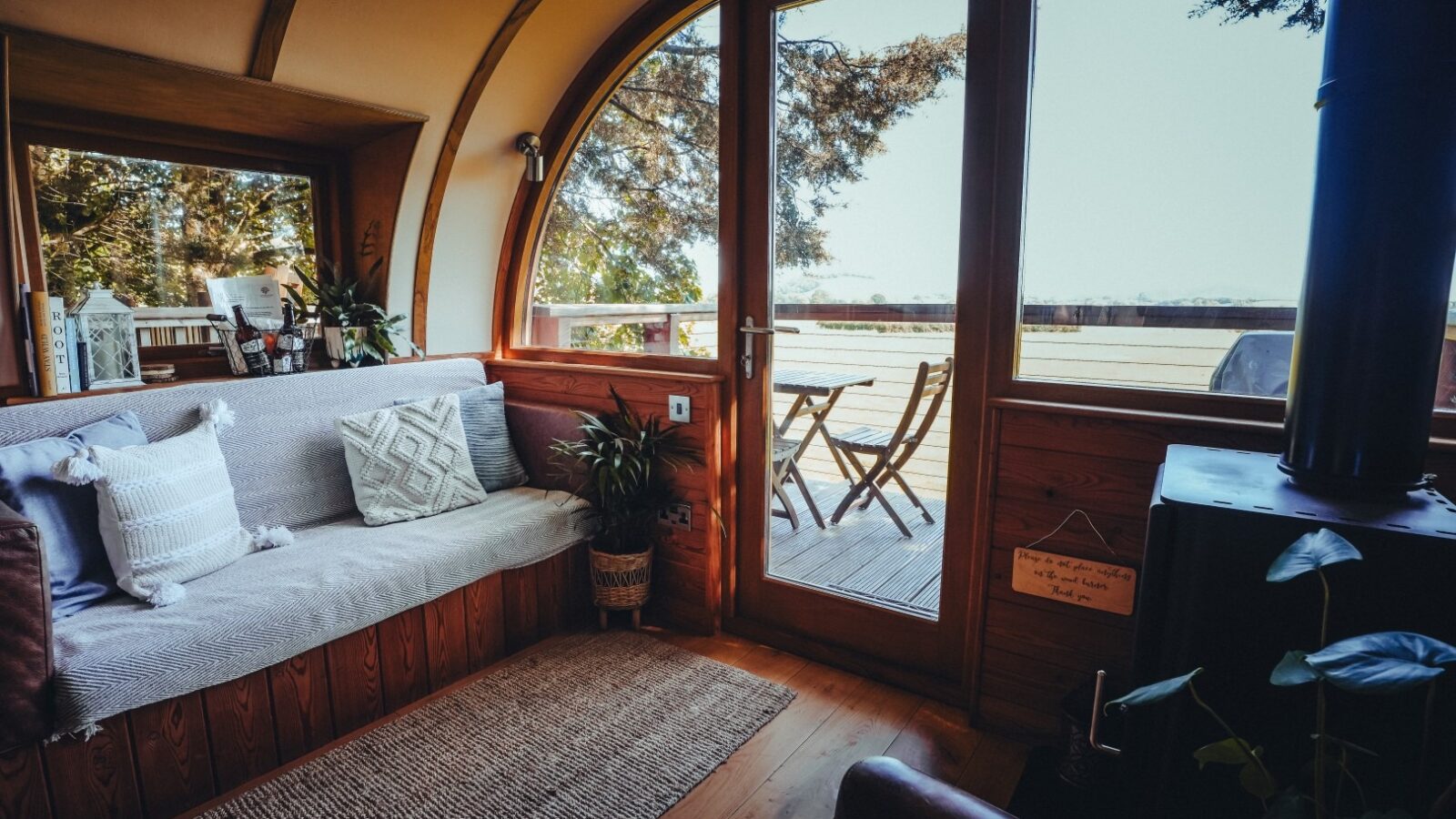 A cozy wooden cabin interior with a large arched window and a glass door. It features a cushioned bench with pillows, several potted plants, and a small wood stove. Outside, the wooden deck at The Orchard overlooks a lush, sunny landscape with views of Fenny Castle in the distance.