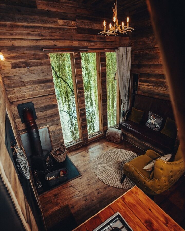 A cozy, rustic living room with wooden walls and floors. It has a small stove, a green armchair, a brown couch with pillows, a round rug, and tall windows with light curtains offering a view of an orchard outside. There is an antler chandelier hanging from the ceiling.