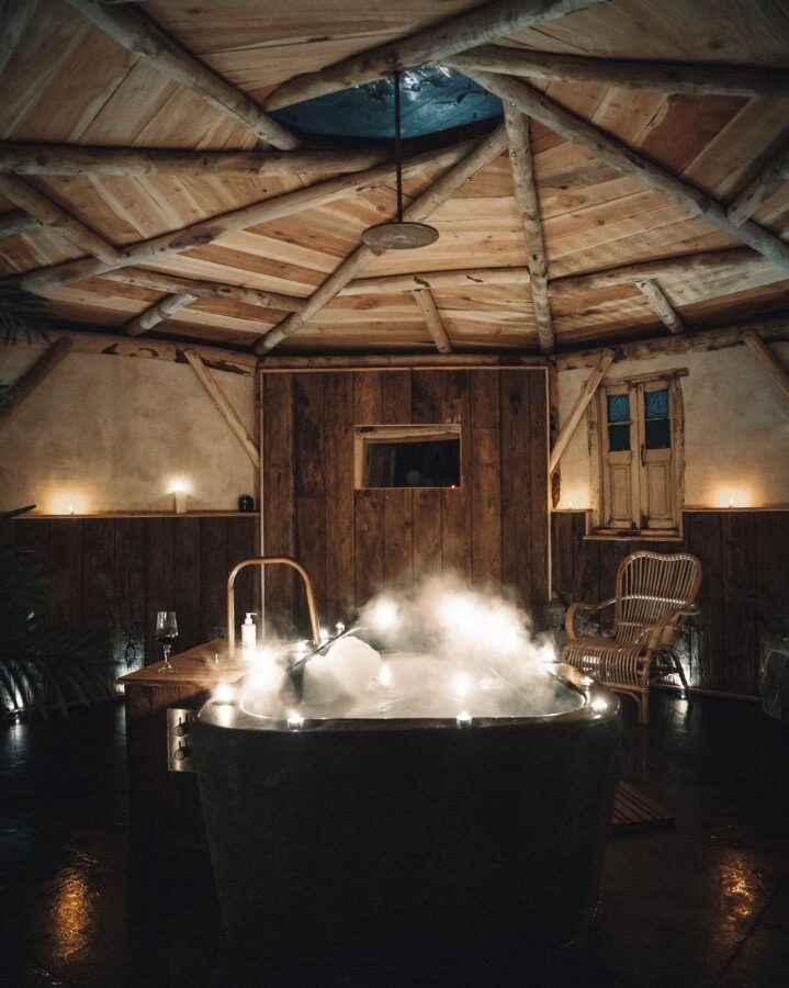 A cozy, rustic room with wooden beams and walls offers the charm of The Orchard. At the center is a large, steaming bathtub surrounded by soft, ambient lighting. There's a wicker chair to the right and a small wooden table with a glass of water and candles to the left.