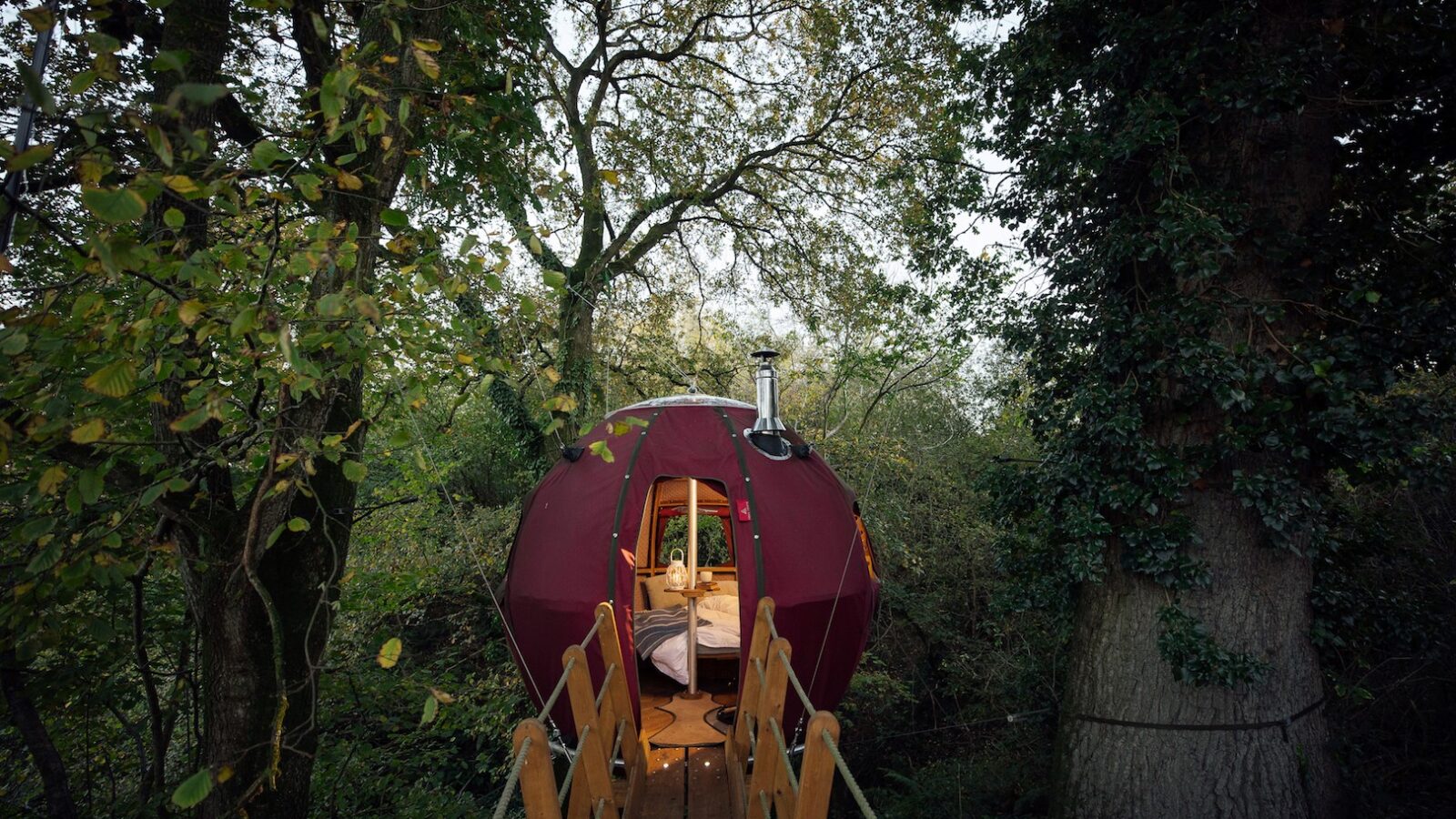 A spherical, maroon-colored treehouse suspended among lush, green trees on Pennard Hill can be accessed via a wooden bridge. Its door is slightly open, revealing a cozy interior with warm lighting and a neatly made bed inside—like one of those unique Tree Tents.