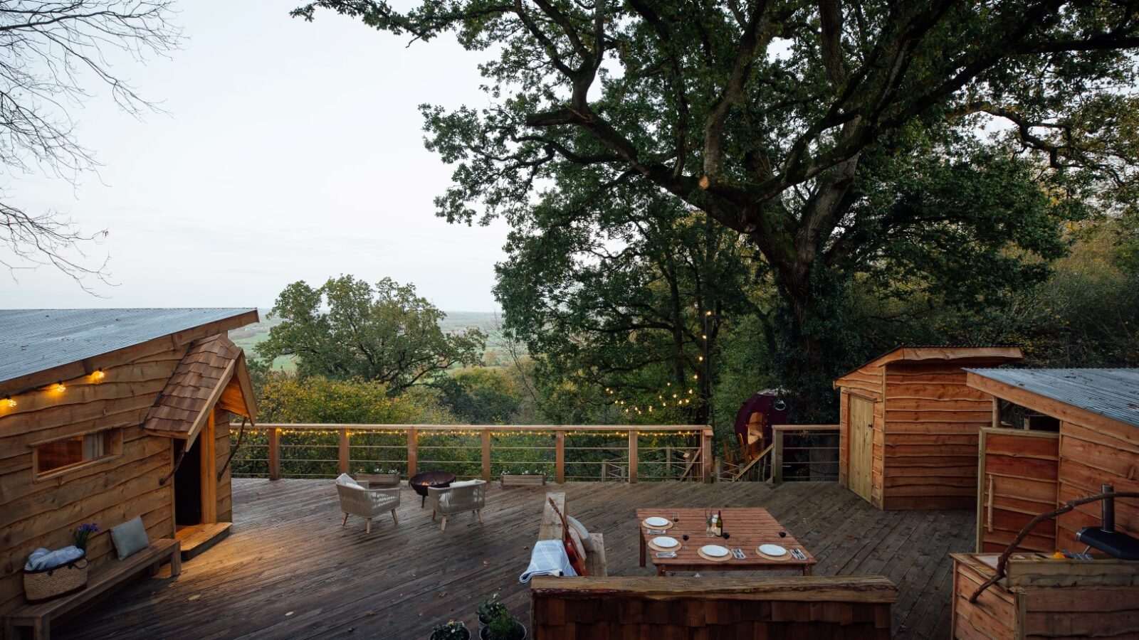 A cozy outdoor wooden deck setup at Pennard Hill, surrounded by tall trees. The deck features a table set for a meal, cushioned seating areas, string lights, and several rustic wooden cabins. The background shows a scenic view of greenery and a distant horizon with tree tents nestled among the branches.