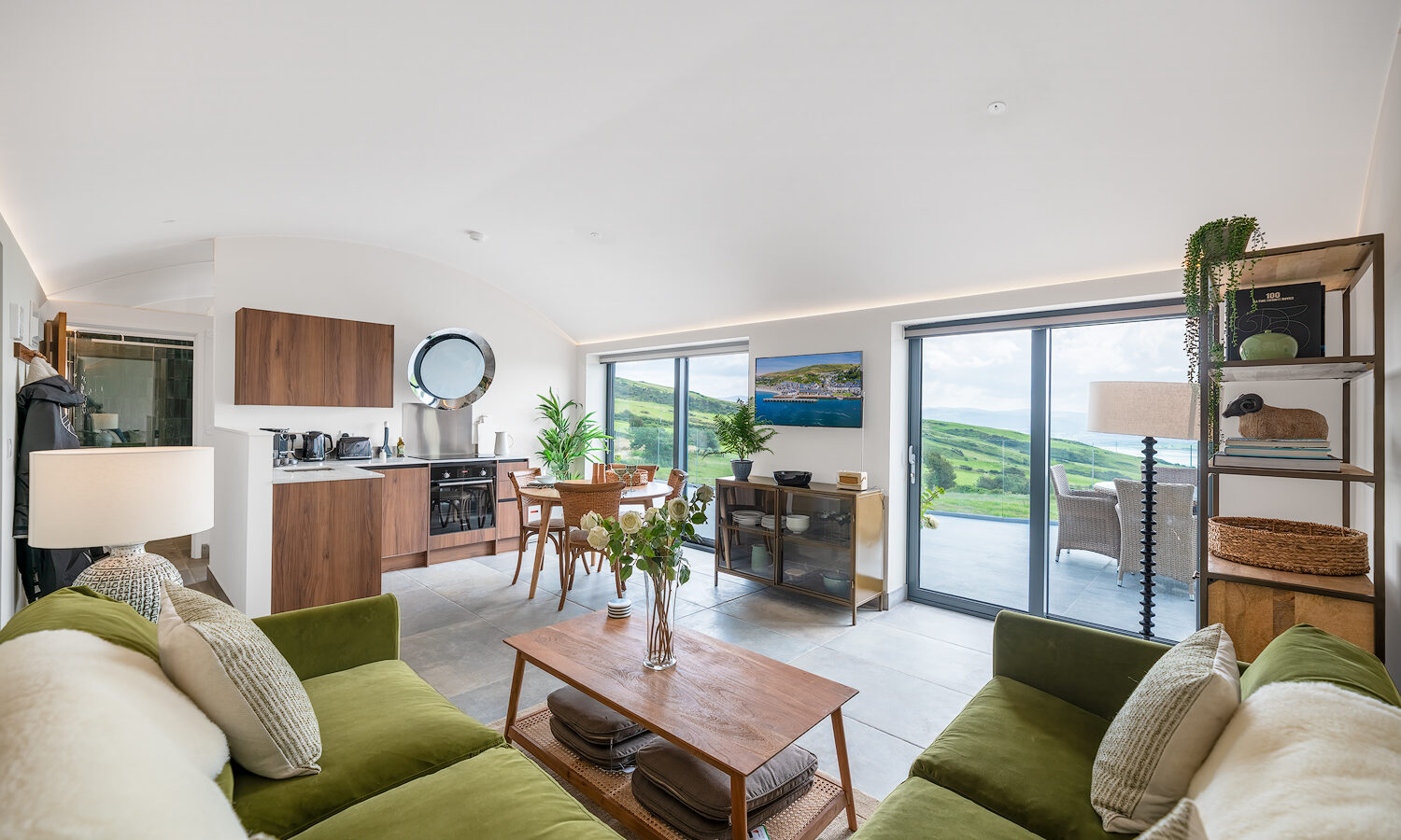 A modern, open-plan living area with a green sofa, wooden coffee table, and indoor plants. A kitchen with wooden cabinetry is visible on the left. Large glass doors open to a scenic view of rolling green hills and a clear sky over Willow Hill. A TV is mounted on the wall.