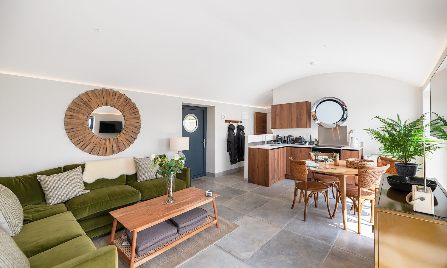 A modern, open-plan living area at The Willow Hill features a green velvet sofa, wooden coffee table, and a circular wall mirror. Adjacent is a kitchen with wooden cabinets, a round window, a dining table with wicker chairs, and a large potted plant. The room is bright and spacious.