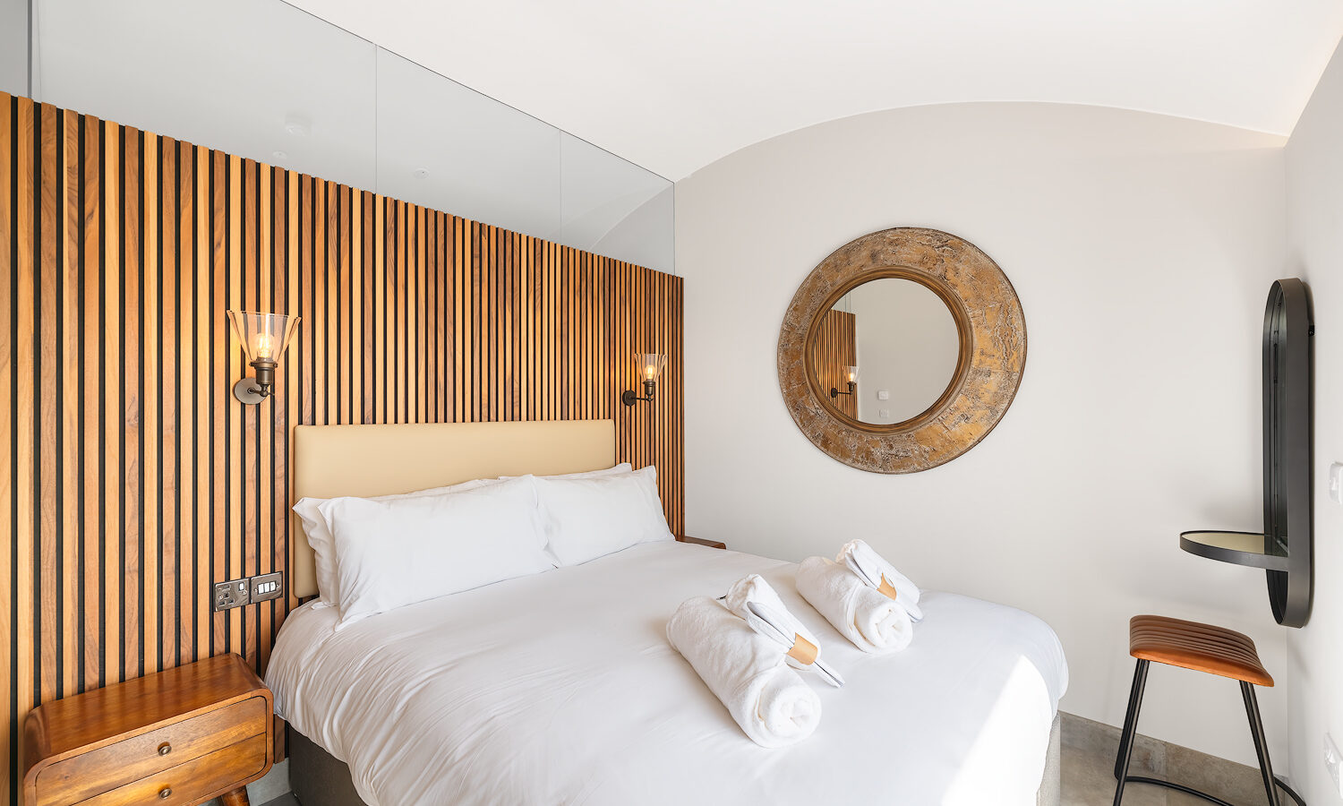 A modern bedroom at The Willow Hill features a cozy bed with white linens and neatly folded towels. The headboard wall is adorned with vertical wooden slats, while two wall-mounted sconces provide ambient lighting. A large round mirror hangs on the adjacent wall above a small side table.