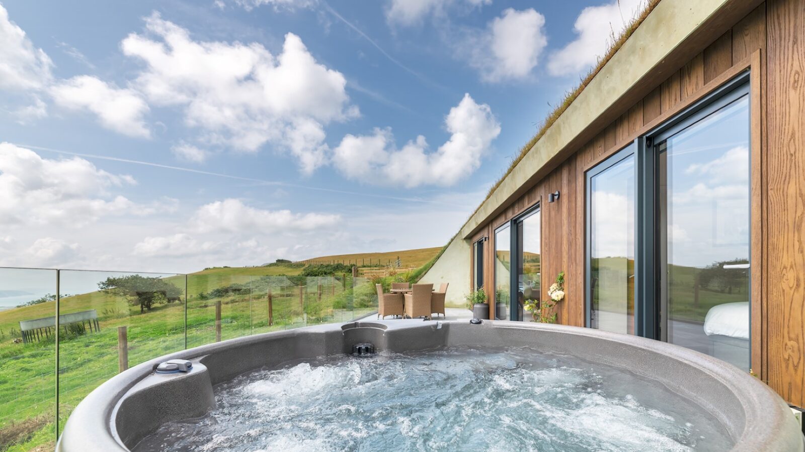 A bubbling outdoor hot tub on a wooden deck at The Willow Hill overlooks rolling green hills and a distant body of water. The deck, adjacent to a modern building with large glass windows, sits under a partly cloudy blue sky. A small table with chairs is visible on the deck.