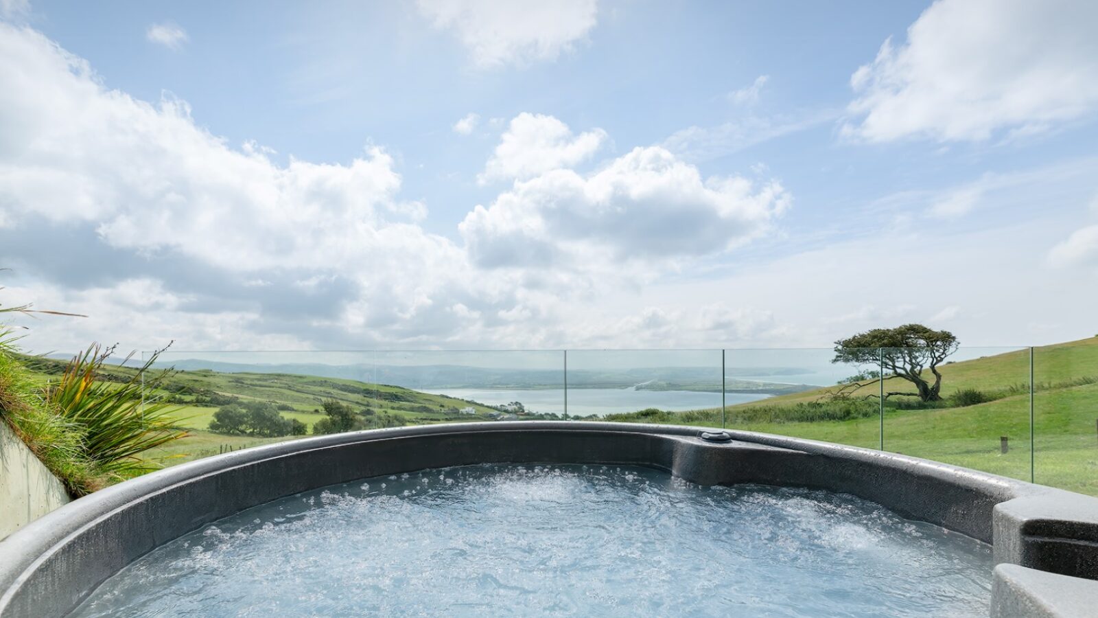 A bubbling outdoor hot tub is set against the scenic view of Willow Hill's rolling green slopes and a distant body of water under a bright, partly cloudy sky. A glass fence surrounds the area, enhancing the open and serene atmosphere.