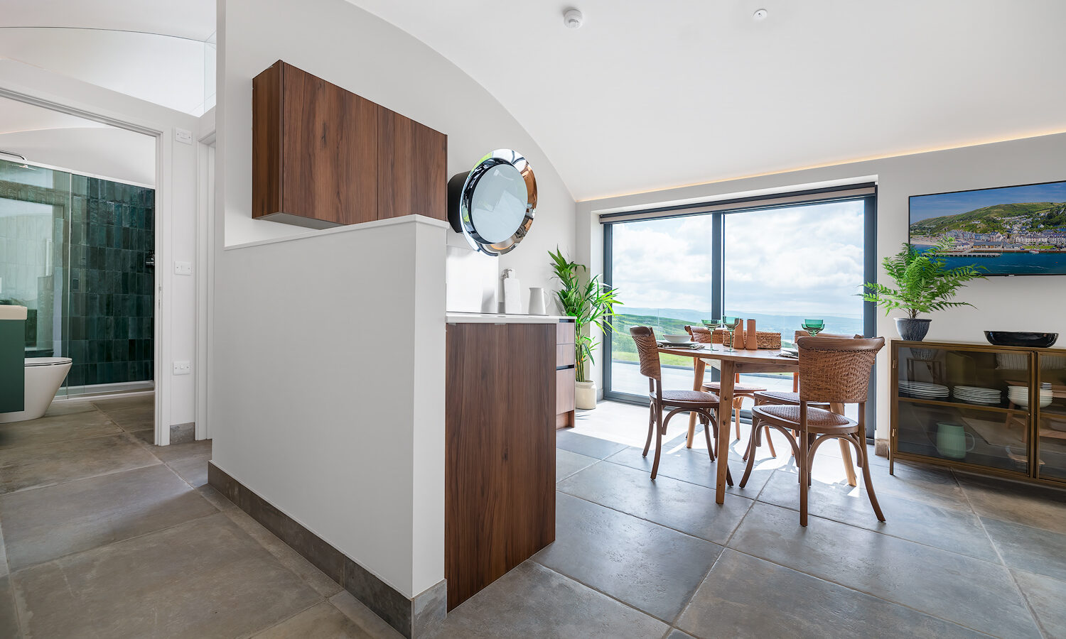 A modern open-plan kitchen and dining area with large sliding glass doors allowing natural light awaits you at Willow Hill lodging. The dining table is set with four chairs, and plants, cabinets, and wall art enhance the decor. A partial view of a bathroom with green tiles is visible to the left.