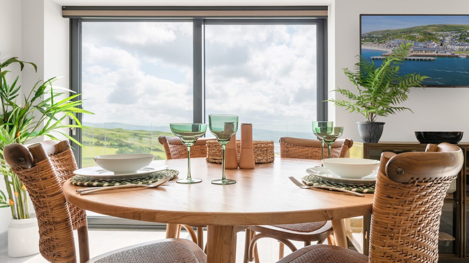 A bright dining area with a round wooden table set for four. The table features green glassware, plates, and napkins. Large windows offer a scenic view of Willow Hill under a partly cloudy sky. Green plants adorn the room, and a colorful painting graces the wall.