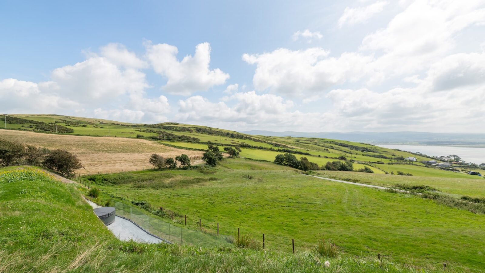 The image showcases a picturesque landscape with rolling green hills under a partly cloudy sky. In the foreground, there is a fenced area and a small path meandering through the grass. The background features vast open fields and the serene waters near Willow Hill.