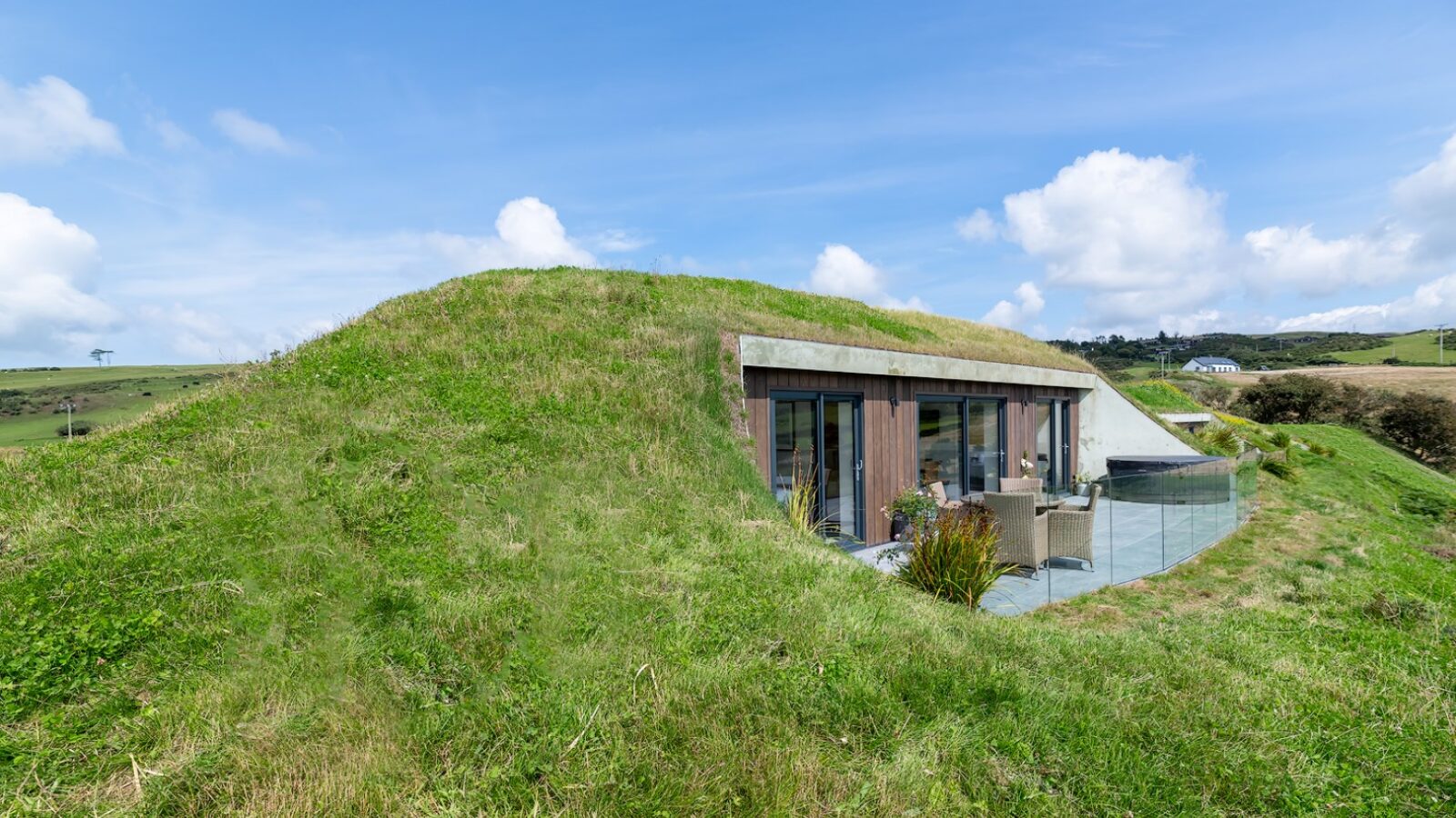 A modern, eco-friendly house, aptly named The Willow Hill, boasts a green, grassy roof and is partially embedded into a hillside. The exterior features large glass windows and a patio area. The surrounding landscape is lush with greenery under a blue sky with scattered clouds.