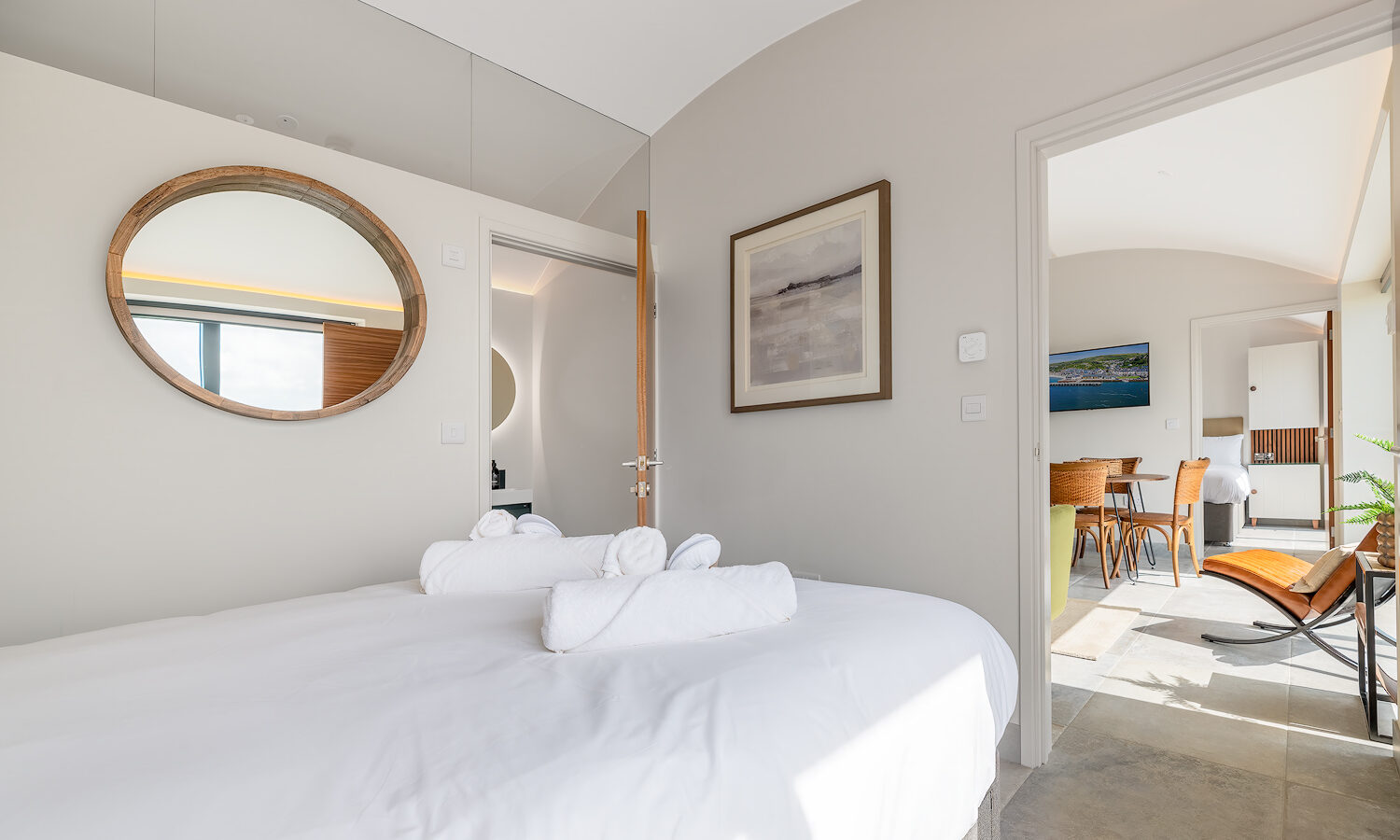 A bright bedroom in The Willow Hill features a neatly made bed with white linens and rolled towels. A circular mirror hangs above the bed, reflecting light from the window. An open door reveals a dining area with wooden furniture, a wall-mounted TV, and a glimpse of contemporary decor.