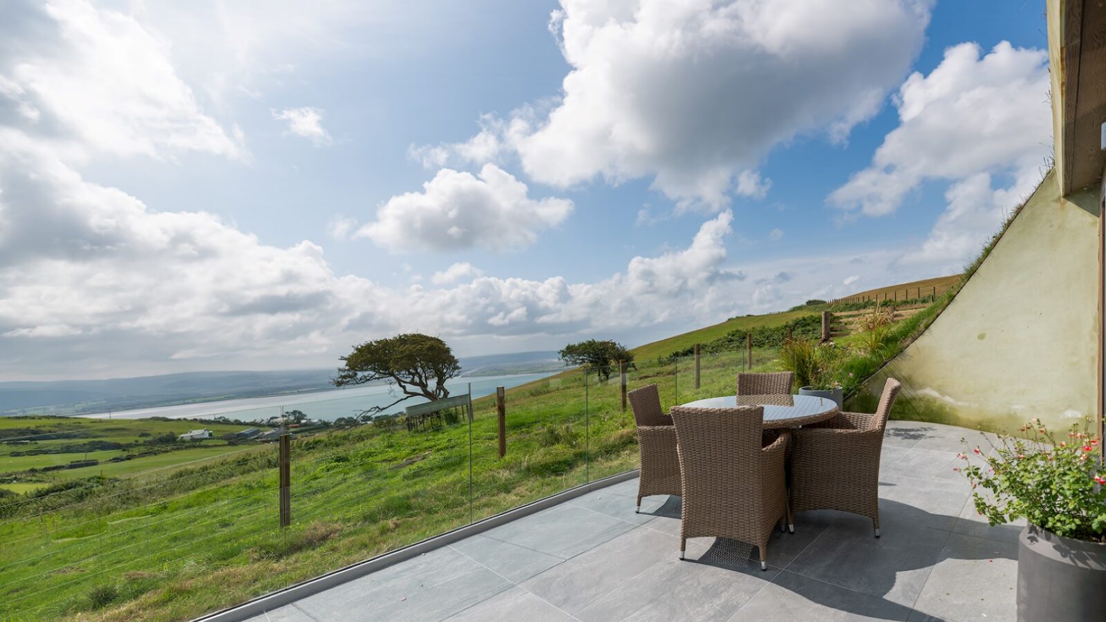 A scenic view of a patio with an outdoor dining set featuring a round glass-top table and four wicker chairs. The patio overlooks Willow Hill's lush green landscape, with rolling hills and a distant body of water under a partly cloudy sky, all bordered by a wooden fence.