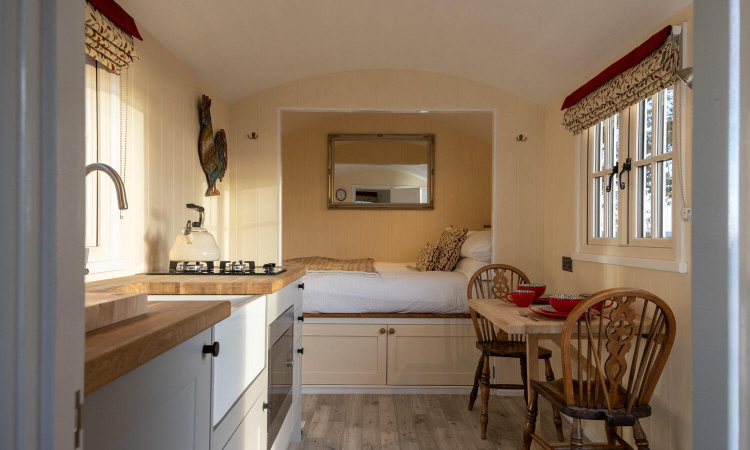 The interior of this cozy Shepherds Hut features a small kitchen with a wooden countertop on the left, a dining area with a table and two chairs on the right, and a bed with storage drawers underneath at the back. Soft lighting and decorative elements from Abbey Farm adorn the space.