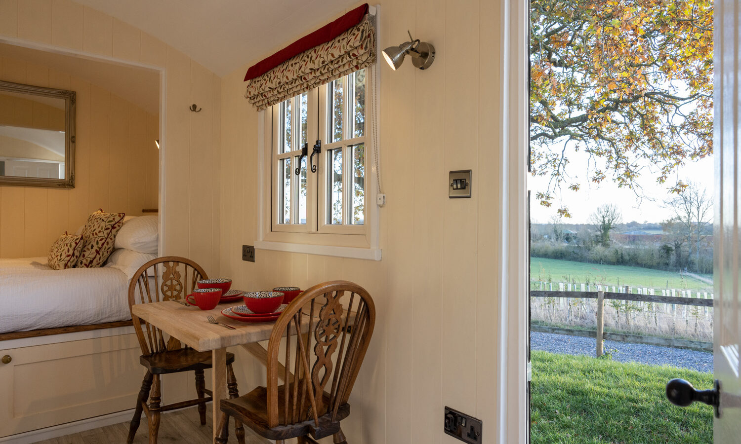 The cozy interior of a small cabin echoes the charm of traditional shepherds huts, with a wooden table set for two and a bed in view. A large window and open door reveal Abbey Farm's scenic landscape, where fields stretch under a clear sky. Natural light enhances the space's warmth and appeal.