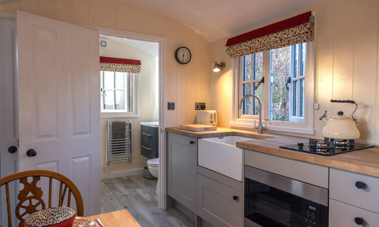 A cozy kitchen at Abbey Farm features wooden cabinets, a farmhouse sink, and a gas stovetop. A clock hangs above the microwave, while a small window with red curtains lets in natural light. In the background, there's a glimpse of the bathroom characteristic of charming farm stays.