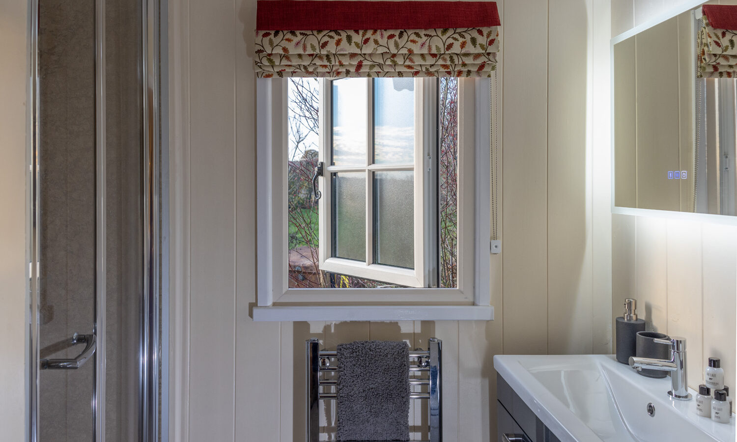 A bathroom with a shower on the left, an open window adorned with a floral-patterned valance reminiscent of Abbey Farm charm, and a sink with a large mirrored cabinet. A towel hangs on a heated rack under the window, and toiletries are neatly arranged on the sink counter.