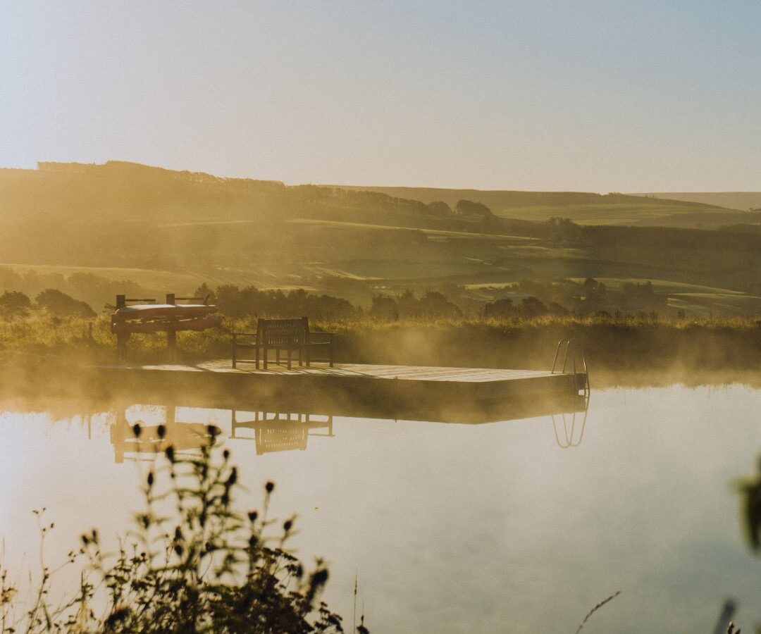 A serene lakeside scene at dawn with a wooden dock extending into the misty water, bordered by grasses and wildflowers softly lit by the rising sun. The dock has a metal ladder and a bench, while tiny homes peek from behind rolling hills under a clear, light-blue sky.