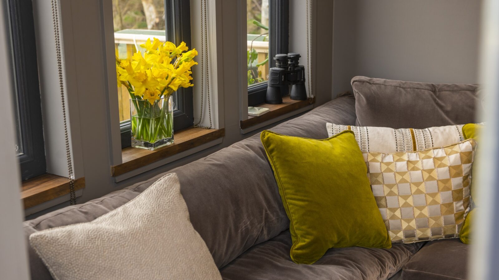 A cozy living room corner in a tiny home features a gray cushioned sofa adorned with yellow and patterned throw pillows. A vase of bright yellow daffodils and a pair of binoculars rest on the wooden window sill, with large windows offering a view beyond the borders of your retreat.